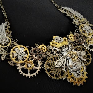 May include: A gold and silver steampunk necklace featuring a bee with intricate wings and gears. The necklace is adorned with floral details and a crown.