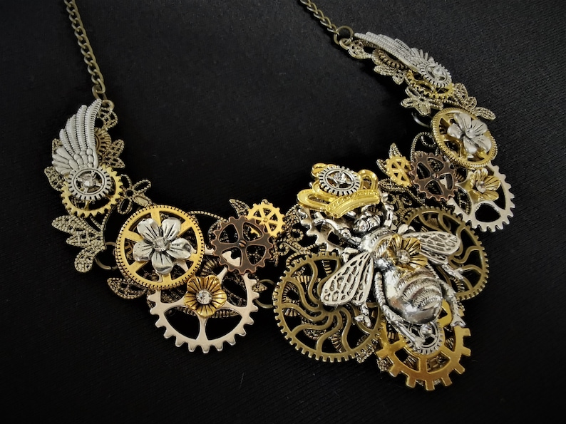 May include: A gold and silver steampunk necklace featuring a bee with intricate wings and gears. The necklace is adorned with floral details and a crown.