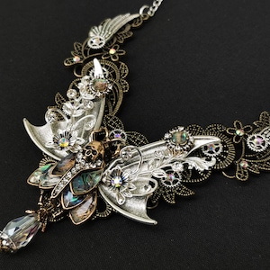 K&ouml;nnte beinhalten: Eine Halskette aus Silber und Bronze mit einem gro&szlig;en, verzierten Schmetterling mit schillernden Fl&uuml;geln und einem Totenkopfdetail. Der Schmetterling ist von filigranen Blumen- und Zahnradmustern umgeben.