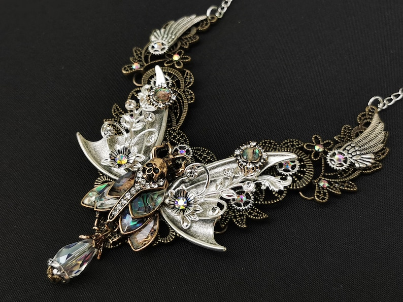 K&ouml;nnte beinhalten: Eine Halskette aus Silber und Bronze mit einem gro&szlig;en, verzierten Schmetterling mit schillernden Fl&uuml;geln und einem Totenkopfdetail. Der Schmetterling ist von filigranen Blumen- und Zahnradmustern umgeben.
