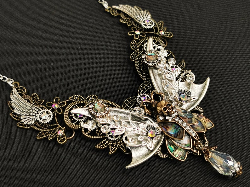 K&ouml;nnte beinhalten: Eine silberne und bronzene Halskette mit einem gro&szlig;en, verzierten Schmetterlingsanh&auml;nger. Der Schmetterling hat schillernde Fl&uuml;gel und einen sch&auml;delartigen Kopf. Die Halskette ist mit kleinen, funkelnden Kristallen verziert.