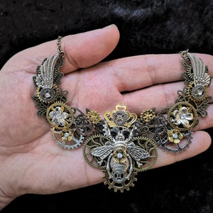 May include: A silver and gold steampunk-style necklace featuring a bee with a crown, gears, and flowers.