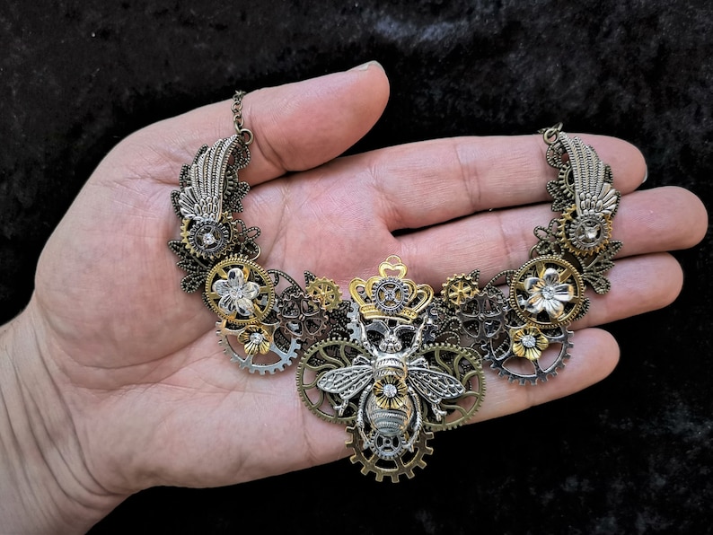 May include: A silver and gold steampunk-style necklace featuring a bee with a crown, gears, and flowers.