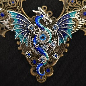 May include: A blue and green dragon with wings made of gears and rhinestones, set against a bronze background. The dragon is a steampunk style pendant.