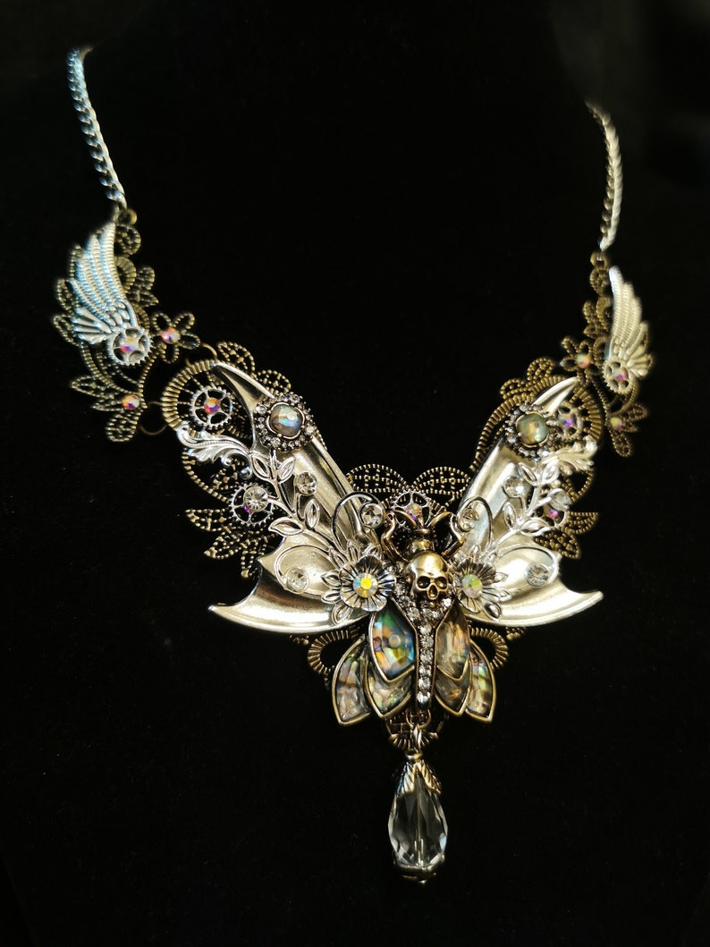 K&ouml;nnte beinhalten: Eine silberne und goldene Halskette mit einem Totenkopf- und Schmetterlingsdesign. Die Halskette ist mit filigranen Details und schillernden Steinen verziert.