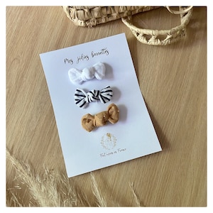 以下が含まれることがあります： 白、青と白のストライプ、白のドット柄の茶色の3つの布製のヘアボウ。ボウは、「Mes jolies barrettes」と「Fait main en France」のテキストが書かれた白いカードに表示されています。