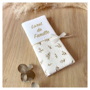 Peut inclure: Un livre en tissu blanc avec une broderie dorée qui dit "Livret de Famille" et un ruban blanc. Le livre est enveloppé dans un tissu blanc avec un motif de feuilles dorées.
