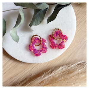 May include: A pair of hoop earrings featuring flower-shaped charms made of translucent pink resin embedded with chunky iridescent glitter. Each charm is suspended from a thin, circular gold-toned metal hoop, creating a bright and reflective accessory.