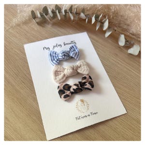 以下が含まれることがあります： 青と白のストライプ、ベージュ、ヒョウ柄の3つの小さなリボンヘアバレッタ。バレッタは、「Mes jolies barrettes」と「Fait main en France」の文字が入った白いカードに配置されています。