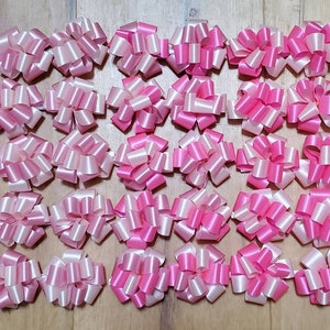 May include: A collection of fifty gift bows arranged in a grid pattern. The bows are made of shiny ribbon in alternating shades of bright pink and light pastel pink, each featuring a multi-looped, circular design for decorative packaging.