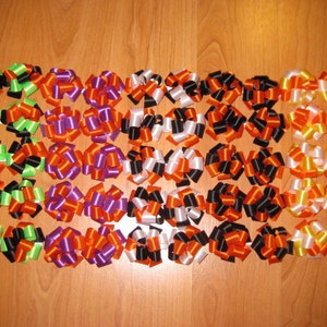 May include: A collection of forty decorative ribbon bows arranged in a grid. The bows feature various color combinations including green and black, purple and orange, black and white, orange and black, and yellow and white satin ribbon loops.