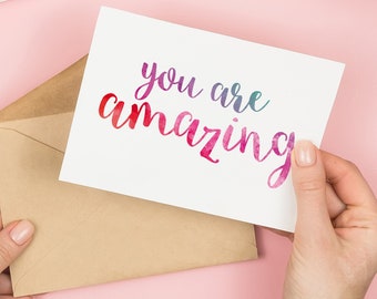 May include: A white card with the text "you are amazing" written in colourful script. The card is being held by a hand and is partially inside a brown envelope.