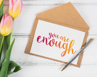 May include: A white card with the text "you are enough" in a colourful gradient font, inside a brown envelope. A bouquet of pink and yellow tulips sits on a white wooden surface next to the envelope.