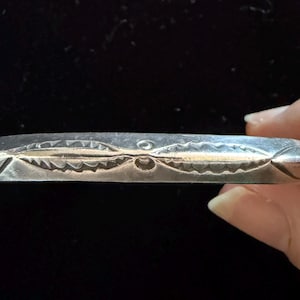 May include: A silver-toned metal bracelet featuring a polished finish and a central horizontal design element. The surface is decorated with two symmetrical, leaf-like engraved patterns separated by a small circular indentation in the middle.