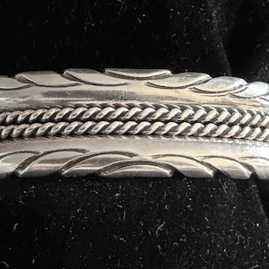 May include: A silver cuff bracelet featuring a central double-twisted rope design flanked by two smooth bands with diagonal etched grooves along the top and bottom edges. The metal has a polished finish with darkened recessed areas for contrast.
