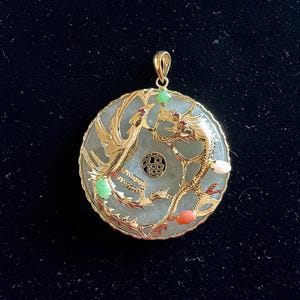 May include: A round, gold-colored pendant with a jade center. The pendant features a dragon and phoenix design with red, green, and white gemstones. The pendant has a gold bail for hanging.