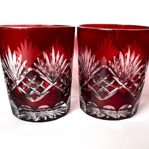 May include: Two matching drinking glasses made of deep red glass with clear cut crystal detailing. The exterior features a repeating pattern of fan-shaped motifs and diamond-shaped cross-hatching, with a scalloped cut design around the base.