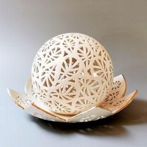 Può includere: Una sfera in ceramica color crema con un motivo geometrico traforato poggia su un piatto a forma di fiore abbinato. Il piatto ha un bordo dorato. La sfera e il piatto sono accessori decorativi per la casa.