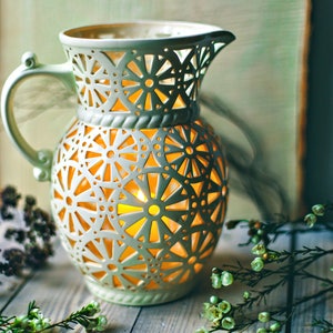 Puede incluir: Una linterna de cerámica blanca con un delicado y complejo diseño de corte. La linterna está iluminada desde el interior, proyectando un cálido resplandor. La linterna está rodeada de ramitas verdes.