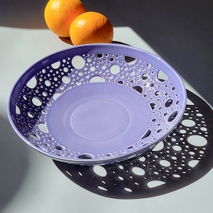 Può includere: Una fruttiera in ceramica lavanda con bordo traforato, con un motivo di fori di varie dimensioni. Due arance sono posizionate sopra la ciotola. L'ombra della ciotola è proiettata sulla superficie sottostante.