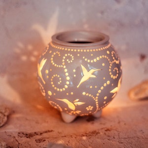 May include: A gray ceramic candle holder with a round shape and three small feet. The holder features cut-out hummingbird and floral designs, with a warm glow emanating from within. The design includes dotted patterns and swirls.