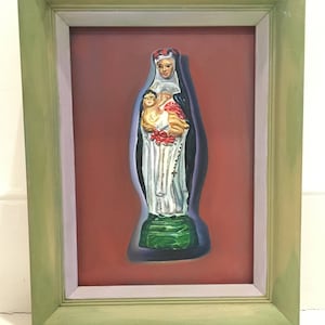 May include: A framed painting of a religious figurine. The figurine is a white and green statue of the Virgin Mary holding a baby Jesus. The statue is set against a red background.