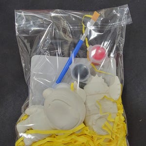May include: A clear plastic bag containing a paint-your-own craft kit. The kit includes unpainted ceramic figurines, a blue-handled paintbrush, paint pots, and yellow paper filler. The figurines are various shapes, including a bear.