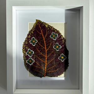 May include: A framed artwork featuring a large, dark burgundy leaf with an orange central vein and dotted pattern. The leaf is adorned with green and white floral designs. The white frame enhances the artwork's presentation, creating a striking visual contrast.