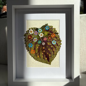 May include: A framed artwork featuring a large, green leaf with painted colorful flower designs. The leaf has a textured surface and is mounted on a light-colored background within a white frame. The flowers are in shades of blue, orange, white, and pink.