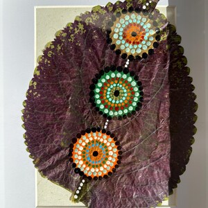 May include: A framed artwork featuring a large, dried leaf with three colorful mandala designs. The leaf is a deep purple with green edges, and the mandalas are painted with orange, blue, green, and black dots. The artwork is displayed in a white frame.
