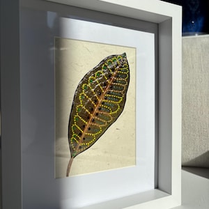 May include: A framed artwork featuring a detailed leaf illustration. The leaf is painted with brown, yellow, and green dots and lines, set against a cream-colored background. The artwork is encased in a white frame, creating a clean, modern aesthetic.