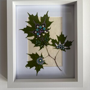 May include: A framed botanical art piece featuring green leaves with intricate dot-painted floral designs in shades of blue, purple, and white. The artwork is set against a neutral background and framed in a white, rectangular frame.
