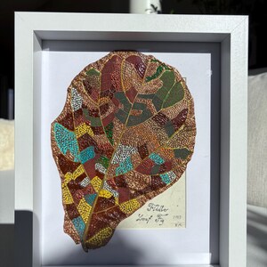 May include: A framed artwork featuring a detailed leaf design. The leaf is composed of various colors, including brown, green, yellow, and blue, arranged in a mosaic pattern. The artwork is set against a white background within a white frame. The text "Fiddle Leaf Fig" is visible.
