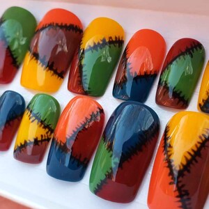 May include: A set of press-on nails with a patchwork design. Each nail features a combination of colors including red, orange, yellow, green, and blue, with black stitching details. The nails are arranged in a white tray.