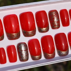 Könnte beinhalten: Ein Set roter, glänzender Kunstnägel. Einige Nägel haben einen metallisch goldenen Sockel, andere ein goldenes Wirbelmuster. Die Nägel werden in einem weißen Tablett präsentiert und sind gebrauchsfertig. Maße nicht verfügbar.