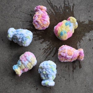 May include: Six colorful crocheted yarn balls in various shades of pink, blue, yellow, and white. The balls are arranged on a gray concrete surface.