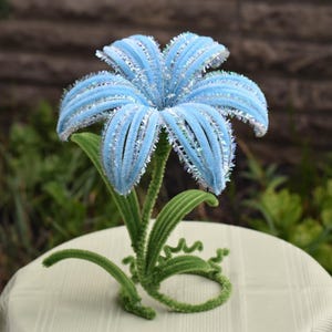 Könnte beinhalten: Eine dekorative, hellblaue Blume mit silbernen Glitzerakzenten. Die Blume hat mehrere Blütenblätter und wird von einem grünen Stiel und Blättern getragen. Die Blume ist etwa 20 cm hoch.
