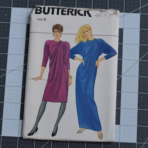 May include: A vintage Butterick sewing pattern, size 8, featuring illustrations of two draped dresses. One dress is a deep magenta, the other is a vibrant blue. The pattern is on a black and gray grid cutting mat.