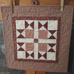May include: A small, square quilt with a star pattern in the center. The quilt features a patchwork design with shades of red, cream, and brown. The quilt is held up by two wooden clothespins.