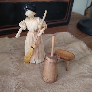 May include: A handmade doll made of straw, holding a broom, next to a wooden butter churn and a small wooden table. The doll has black hair and is wearing a light-colored dress. The butter churn and table are made of light brown wood.