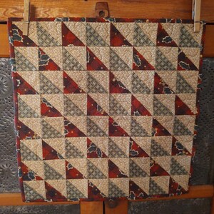 May include: A square quilted wall hanging featuring a repeating half-square triangle pattern. The fabric design alternates between a tan speckled print, a dark red holly leaf pattern, and a muted green geometric print with visible stitching.