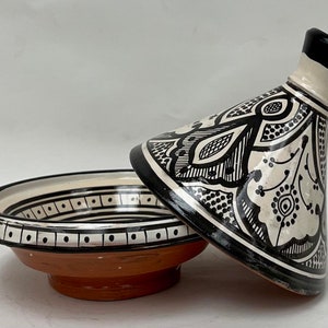 May include: A white ceramic tagine with a black and white geometric design. The tagine has a round base and a conical lid with a knob on top.
