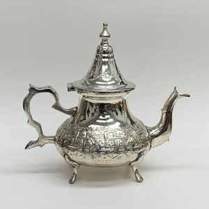 Peut inclure: Une théière en argent avec un design orné et un couvercle. La théière a un corps rond et un bec verseur sur le côté. Elle a trois pieds et une poignée.