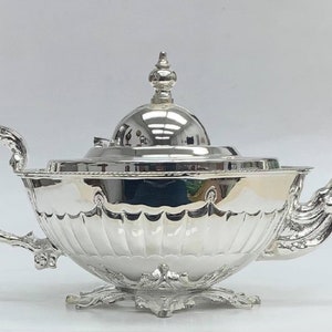 May include: A silver teapot with a decorative handle and spout. The teapot has a domed lid with a finial on top. The teapot is sitting on a decorative base.