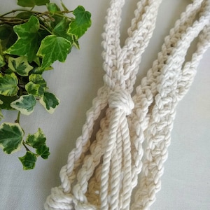 Könnte beinhalten: Ein weißer Makramee-Pflanzenhänger aus Baumwollseil mit einem geknoteten Design.
