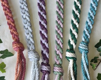 Op de afbeelding: Close-up van zes gevlochten macramé plantenhangers in verschillende kleuren. De kleuren zijn onder meer tinten roze, grijs, paars, groen en blauw. Elke hanger is aan de onderkant geknoopt. De macramé plantenhangers zijn een decoratief item.