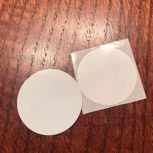 Op de afbeelding: Twee ronde witte stickers op een houten oppervlak. De stickers zijn blanco en hebben een glanzende afwerking.
