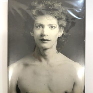 Peut inclure: Portrait en noir et blanc d'une personne aux cheveux bouclés et torse nu. L'image est enfermée dans du plastique transparent, créant une surface réfléchissante. La personne regarde directement le spectateur.