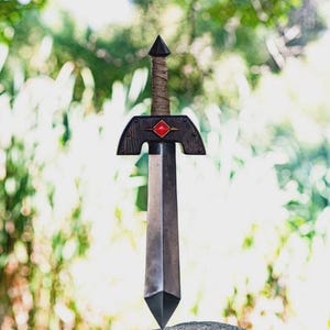 May include: A sword with a dark metallic blade and a pointed tip. The crossguard is dark wood with a central red diamond-shaped inlay. The handle is wrapped in tan cord, topped with a dark pointed pommel. The sword is balanced on a grey stone.