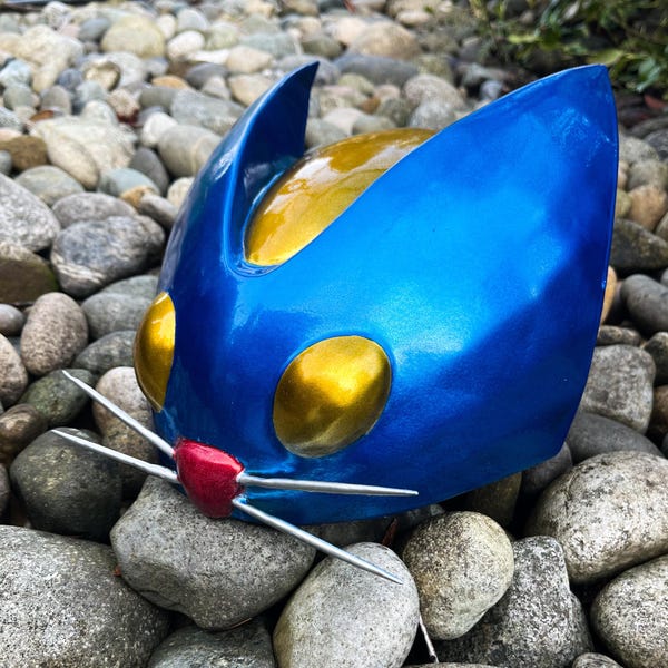 May include: A blue and gold cat-eared helmet with a red nose and silver whiskers. The helmet has large, round, gold-colored eye openings and is set against a background of gray rocks.